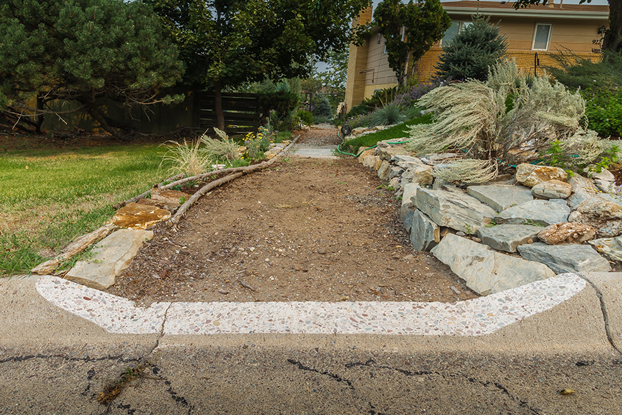 A curb is cut out to make a zero-step wheelchair accessible path leading toward a home. The path's edges are lined with rocks and decorative tree-branches.